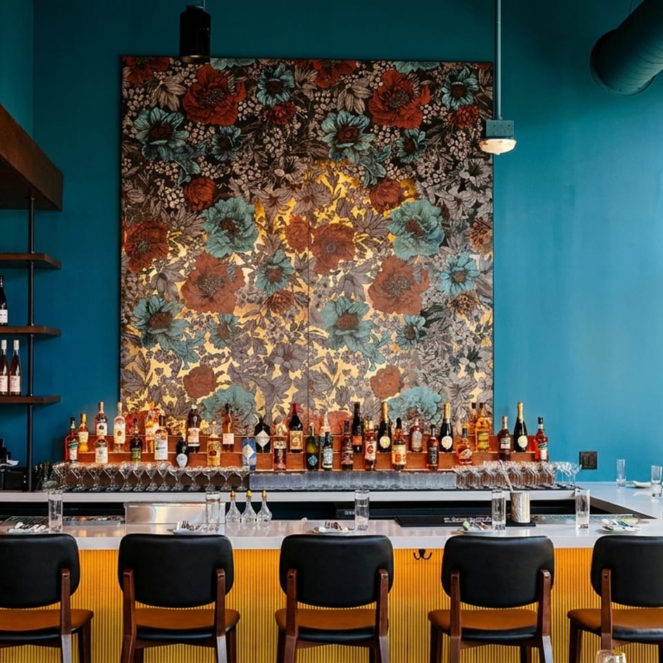 A stylish bar with a teal wall, floral art, bottles, and bar stools.