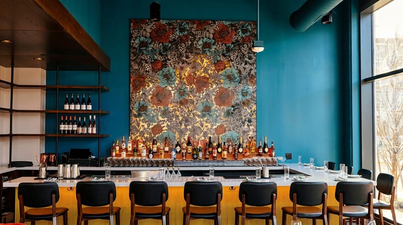 A stylish bar with a teal wall, floral art, and bottles of liquor and wine.