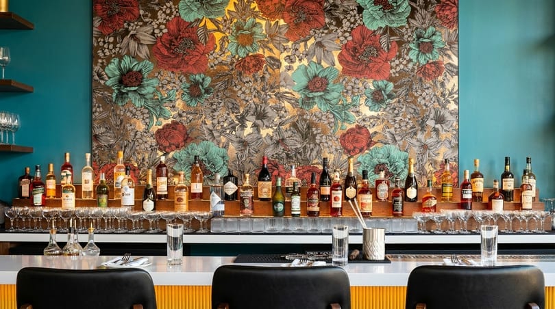 A bar with a floral wallpaper backdrop, displaying various liquor bottles and empty glasses.
