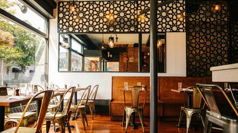 Restaurant interior with tables, gold chairs, large windows, and decorative geometric screens.